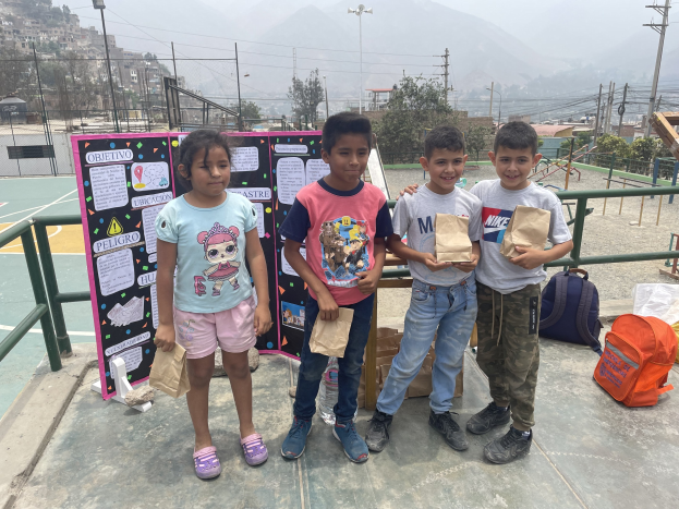 Gruppe von Kindern auf einem Basketballfeld mit Papierbeuteln in den Händen, mit Beuteln und Gegenständen rechts und einer Tafel mit Text links, vor einer Outdoor-Bühne mit Zaun, Pfosten, Straßenlaternen, Bäumen, Gebäuden, Hügeln und Himmel.