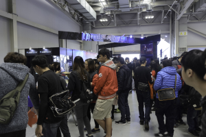 Eine Gruppe von Menschen vor einem vollen Gaming-Expo, mit einigen Besuchern, die Taschen tragen, Texttafeln, Deckenlichter und Rohre im Hintergrund.