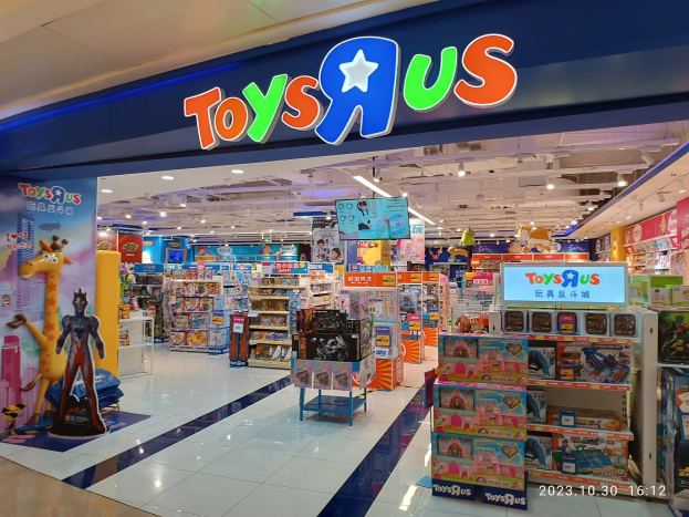 Ein Toys R Us Geschäft in einem Einkaufszentrum, das Regale mit Spielzeug und anderen Artikeln zeigt, ein sichtbares Schild, Deckenbeleuchtung und ein Wasserzeichen in der rechten unteren Ecke.