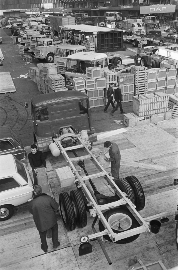 Schwarzes und weißes Foto von Männern, die an einem Truck in einer Fabrik arbeiten, mit Fahrzeugen, Menschen, Kisten und Gegenständen im Hintergrund.