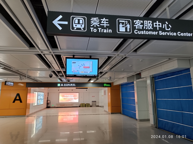 Innenansicht eines Bahnhofs mit einem "Zum Bahnkunden-Service-Zentrum"-Schild oben, sichtbarer Boden, Türen auf der rechten Seite, Deckenanzeigen, einem zentralen Bildschirm und einem Wasserzeichen unten rechts.