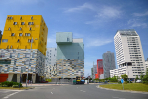 Eine Stadtstraße mit hohen Gebäuden im Hintergrund, Straßenlaternen, Autos auf der Straße, Informationsschilder, Büsche, Bäume und eine bewölkte Himmel, mit der einzigartigen Architektur der Universität Rotterdam.