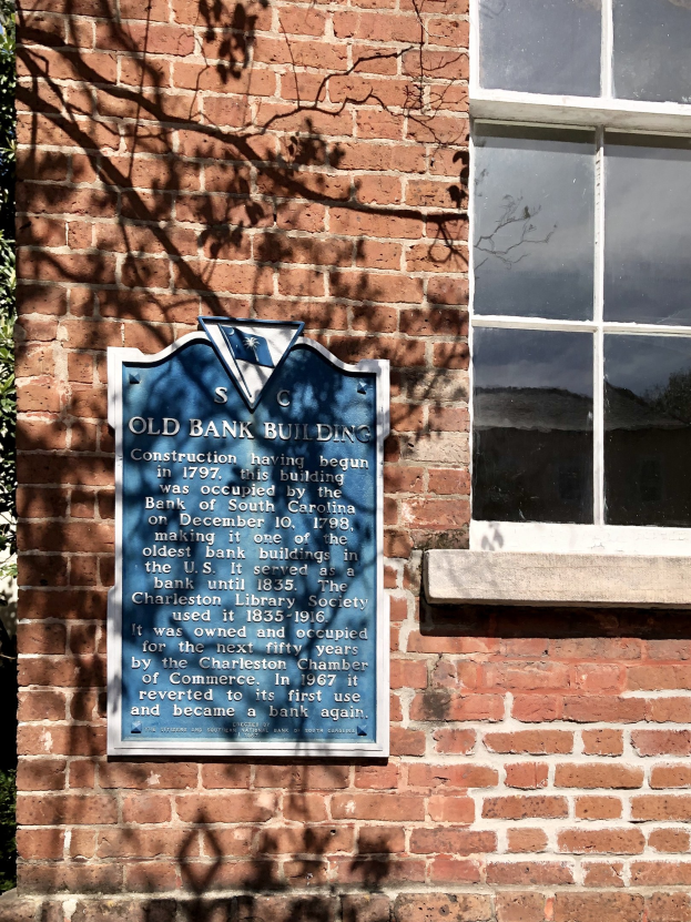 Altes Bankgebäude mit einer blauen Plakette an der Seite, einem Fenster auf der rechten Seite und einem Baum auf der linken Seite.