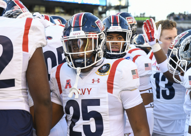 Gruppe von Männern in Fußballausrüstung auf einem Feld mit einer unscharfen Menge, Anzeigetafel mit 'Auburn vs Virginia Tech - 11/28/15 College Football Pick, Odds, and Prediction' und Himmel im Hintergrund.