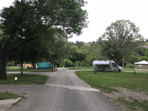Ein Campingplatz mit einem geparkten Auto am Stra├čenrand, Zelten, B├Ąnken und anderen Gegenst├Ąnden auf gr├╝nem Boden, mit B├Ąumen, Geb├Ąuden und einem klaren blauen Himmel im Hintergrund.