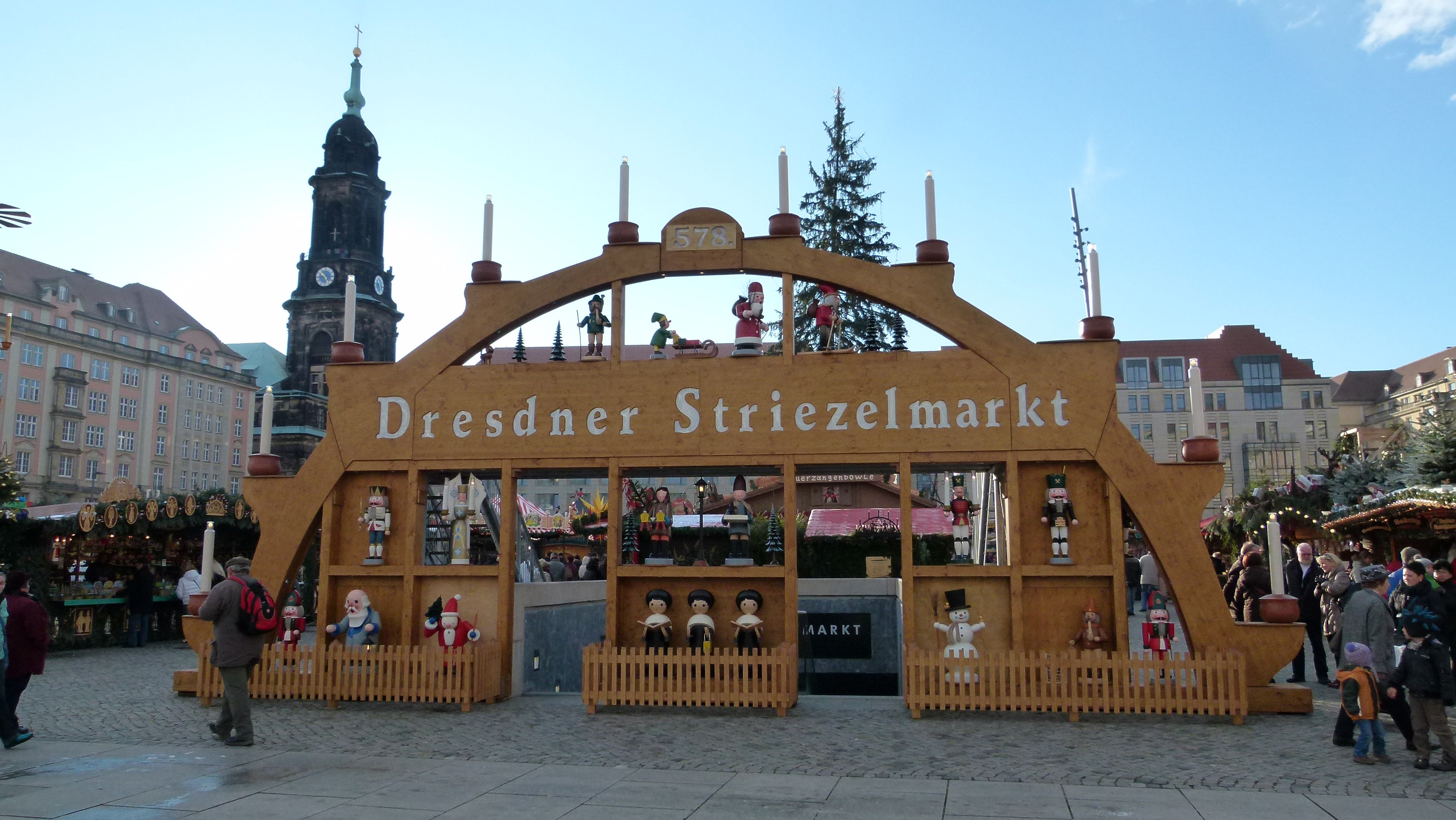 Ein geschäftiges Weihnachtsmarkt in Dresden, Deutschland, mit Menschen, die gehen, Taschen tragen und ein Tor im Hintergrund, umgeben von Bäumen, Gebäuden mit Fenstern und einem bewölktem Himmel.