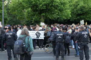 Große Gruppe von Menschen an einer Straße mit Protestschildern, einige tragen Mützen und Taschen, ein Fahrrad im Vordergrund und Bäume und ein Mast im Hintergrund.