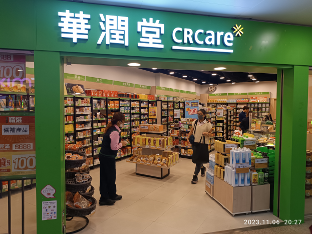 Eine Gruppe von Menschen steht vor einem Geschäft mit einem "crcare"-Schild, in dem sich Regale mit Lebensmitteln, Flaschen und Deckenlampen sowie eine Tafel auf der linken Seite befinden, die darauf hinweist, dass es Teil einer chinesischen Supermarktkette ist, die ihren ersten Online-Shop eröffnet.