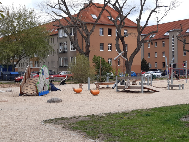 Ein Spielplatz mit einer Rutsche, Schaukeln und anderen Geräten, umgeben von Gras und Bäumen, mit Gebäuden, Fahrzeugen und einem bewölkten Himmel im Hintergrund.