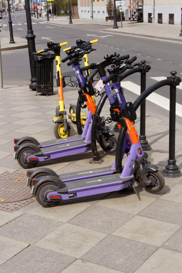 Eine Reihe von Elektro-Scootern, die auf der Straße geparkt sind, mit einem Mülleimer im Vordergrund und Gebäuden, Bäumen, Pfählen, Schildern und Fahrzeugen im Hintergrund.