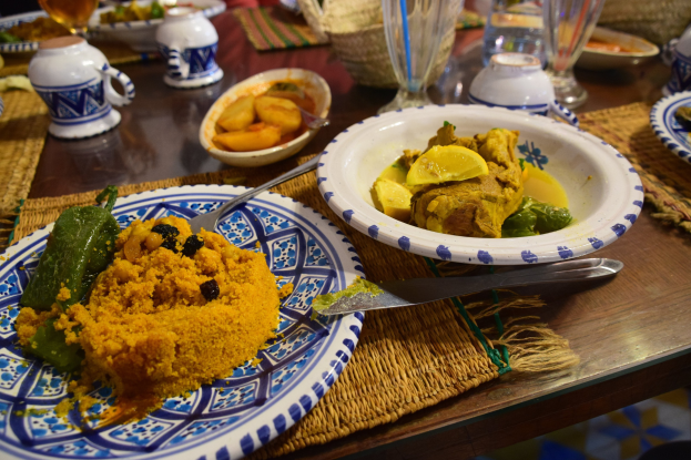 Ein Tisch mit Tellern, Gläsern, Schüsseln, Krügen, Besteck und Matten, umgeben von Menschen im Hintergrund bei einem traditionellen marokkanischen Essen.
