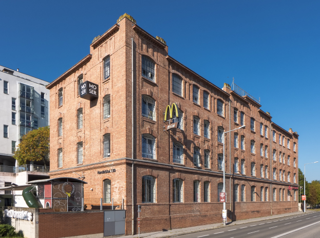 Ein großes McDonald's-Restaurant in einem Backsteingebäude an einer Straßenecke in Berlin, Deutschland, mit umgebender Straßeninfrastruktur, Fahrzeugen, Grünflächen und einem bewölkten Himmel.