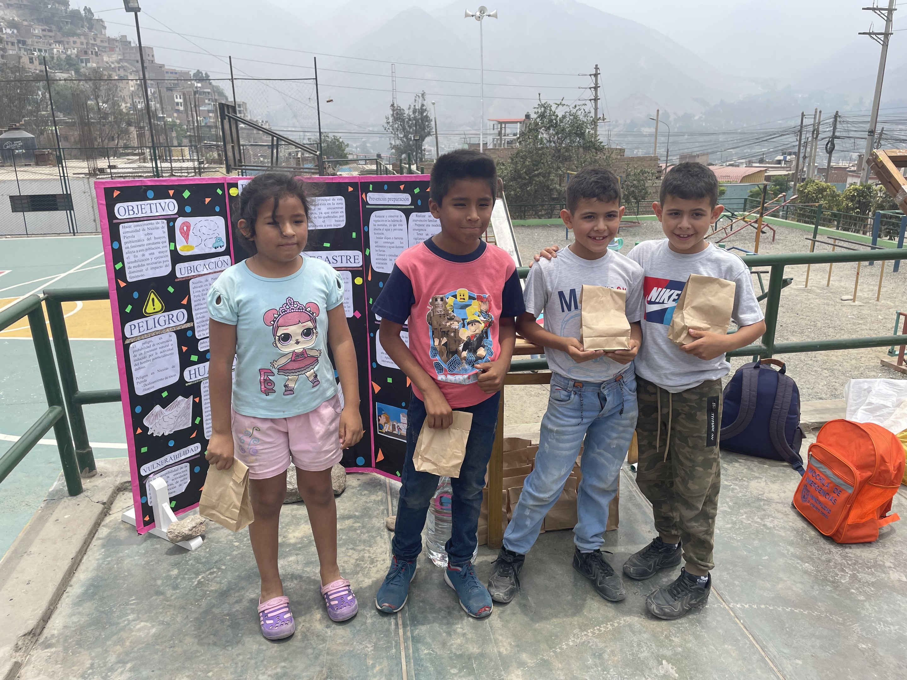 Eine Gruppe von Kindern steht mit Papiertüten auf einem Basketballfeld, rechts davon sind Tüten und Gegenst├Ąnde auf dem Boden und links davon eine Tafel mit Text. Im Hintergrund gibt es Z├Ąune, Masten, Stra├čenlaternen, Strommasten, B├Ąume, Geb├Ąude, H├Ąnge und den Himmel.
