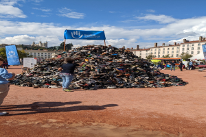 Eine Gruppe von Menschen steht um einen Haufen Schuhe auf einem Feld mit Gebäuden, Bäumen und einem bewölkten Himmel im Hintergrund auf einer internationalen Schuh-Recycling-Veranstaltung.
