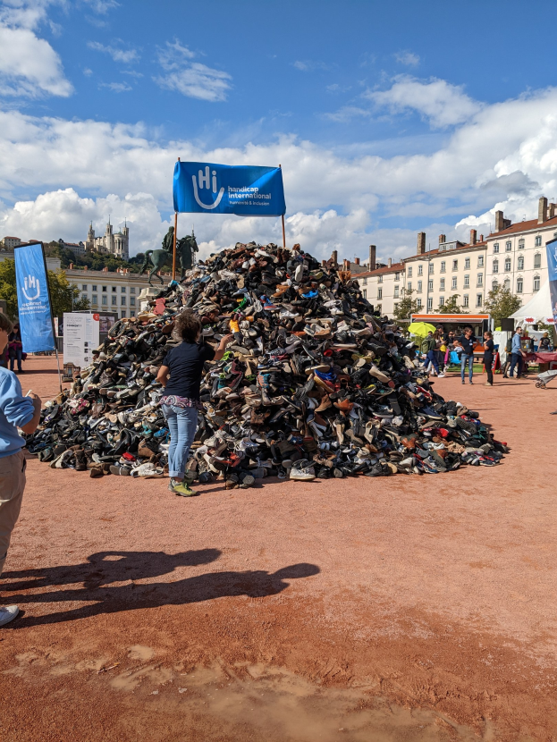 Eine Gruppe von Menschen steht um einen Haufen Schuhe auf einem Feld mit Gebäuden, Bäumen und einem bewölkten Himmel im Hintergrund auf einer internationalen Schuh-Recycling-Veranstaltung.