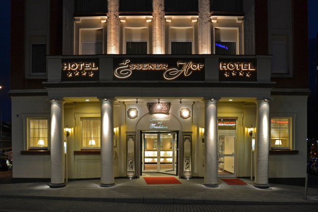 Das Essener Hof Hotel in Berlin, Deutschland, mit seinen Fenstern, Türen, Säulen, Leuchten und Namensschild, das sich entlang einer Straße mit Fahrzeugen auf der rechten Seite und dem Himmel im Hintergrund befindet.