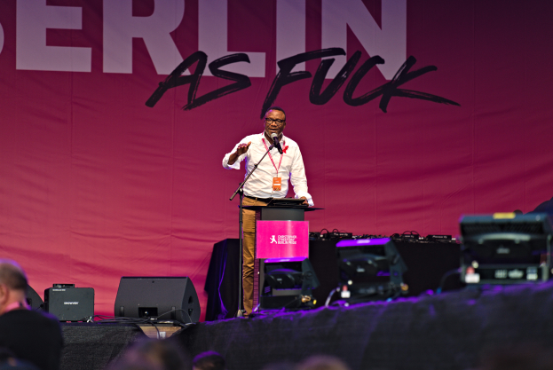 Mann in weißem Hemd und braunen Hosen steht auf einer Bühne und hält ein Mikrofon, mit einem pinken Banner dahinter, das 'Berlin as Fuck' lautet und Lautsprecher auf der Bühne.