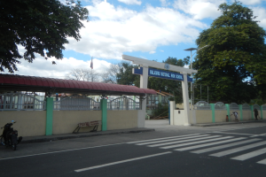 Schuleingang mit einem vor der Tür geparkten Motorrad, eine Mauer mit einem Tor und einem Schuppen, ein Schild, Straßenmarkierungen einschließlich einer Zebrastreifen, Laternenmasten, Bäume, eine Fahne auf einem Mast und ein bewölkter Himmel.