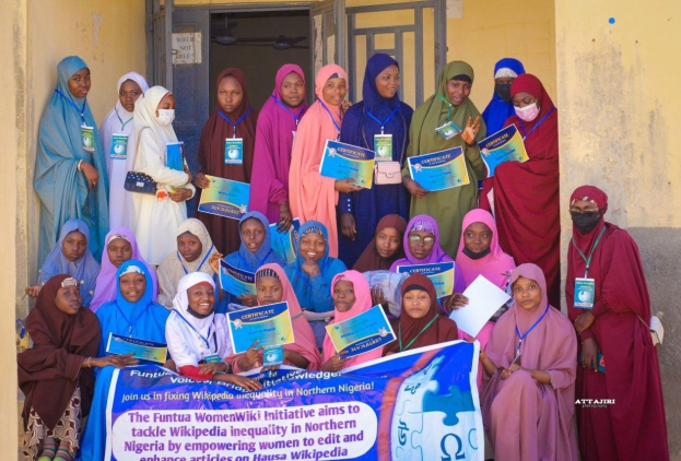 Gruppe von Frauen, die strahlend in die Kamera lächeln und Urkunden in den Händen halten, vor einem Gebäude mit einem Banner, auf dem "Funtua Women Wiki Initiative" steht, im Vordergrund.
