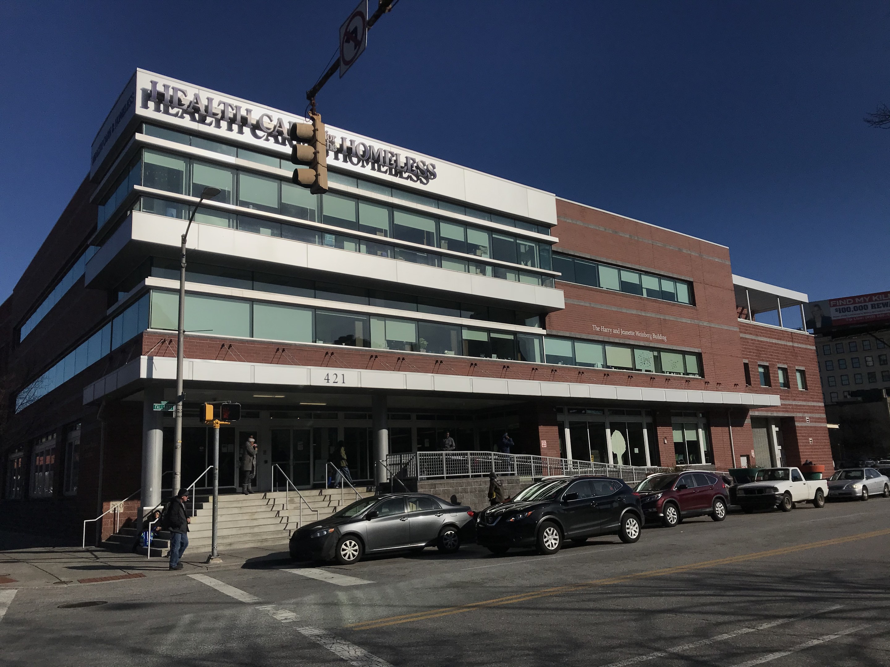Großes Gebäude mit der Aufschrift "New York City Health Care Homeless Center" mit parkenden Autos davor, umgeben von Straßeninfrastruktur, Fußgängern und Bäumen unter einem klaren blauen Himmel.