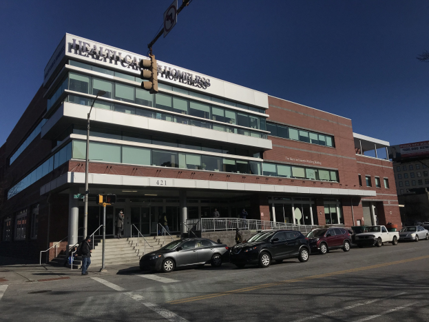 Großes Gebäude mit der Aufschrift "New York City Health Care Homeless Center" mit parkenden Autos davor, umgeben von Straßeninfrastruktur, Fußgängern und Bäumen unter einem klaren blauen Himmel.