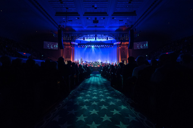 Ein großer Zuschauerraum mit Sitzplätzen, eine Bühne mit Musikern und Überkopfbeleuchtung und -bildschirme, was auf einen Konzertsaal während der Nationalkonvention 2019 hindeutet.