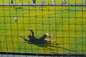 Ein Fussballer hält den Ball vor einem Tor, mit einem Netz im Vordergrund und einigen Menschen im Hintergrund, während mehrere Bälle auf dem Boden verstreut sind.