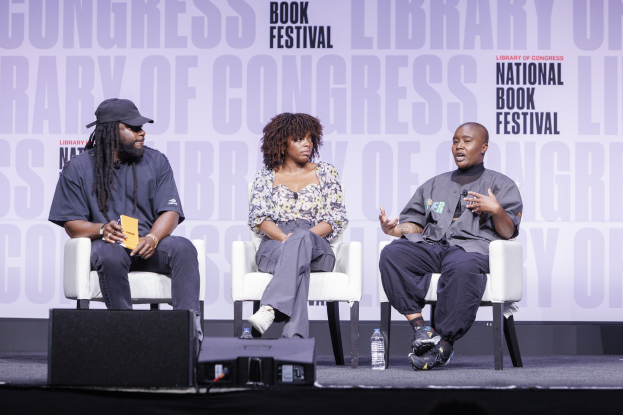 Drei Personen sitzen auf der Bühne bei der Library of Congress Book Festival, mit einem Mann in einer Mütze, der links ein Buch hält, und einer Frau in der Mitte, die ein Mikrofon hält.