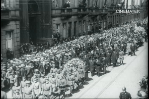 Schwarzes Foto einer Parade mit einer großen Menge, die eine Straße entlangmarschiert, einige halten Gewehre in den Händen, vor einem Gebäude mit einem Wasserzeichen in der rechten oberen Ecke.