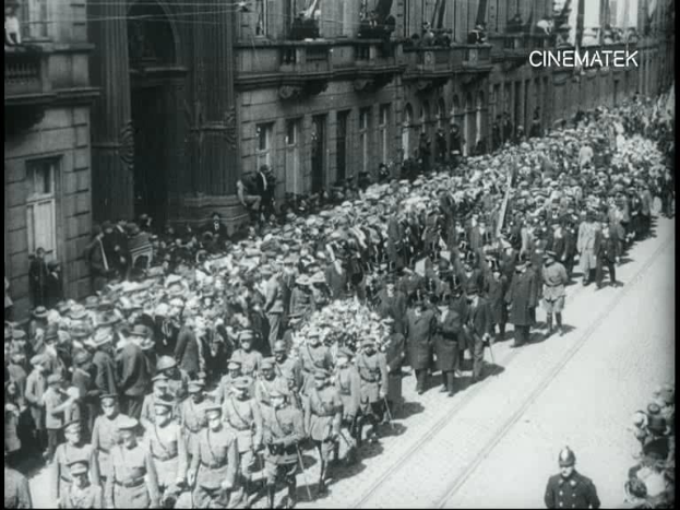 Schwarzes Foto einer Parade mit einer großen Menge, die eine Straße entlangmarschiert, einige halten Gewehre in den Händen, vor einem Gebäude mit einem Wasserzeichen in der rechten oberen Ecke.
