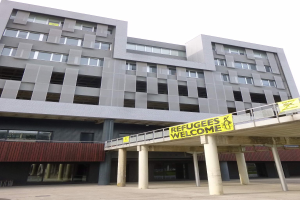 Großes Gebäude mit einem "Geflüchtete Willkommen"-Schild, umgeben von Säulen und einem Geländer, unter einem Himmel-Hintergrund.