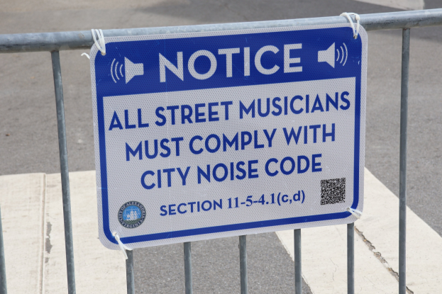 Anschlagtafel an einem Zaun mit einem Schild, auf dem "Anschlag: Alle Straßenmusiker müssen der Stadtlärmverordnung Folge leisten" steht und eine Straße im Hintergrund.