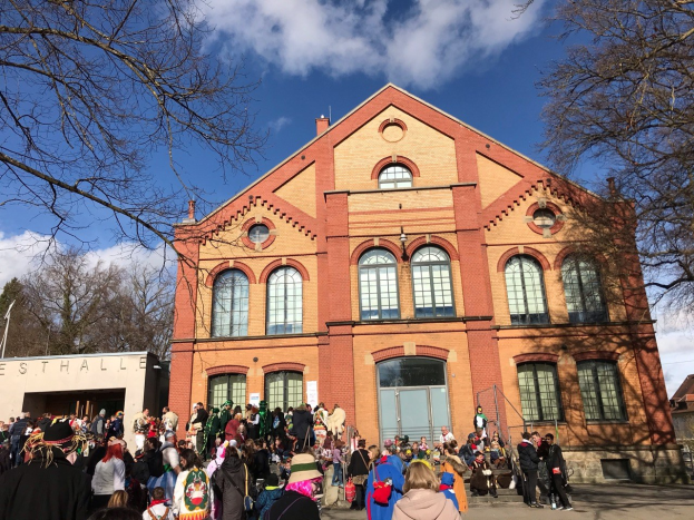 Eine große Gruppe von Menschen, die vor einem mehrstöckigen Gebäude mit Fenstern und Türen stehen, umgeben von Bäumen und einem bewölkten Himmel, einige tragen Mützen und tragen Taschen.