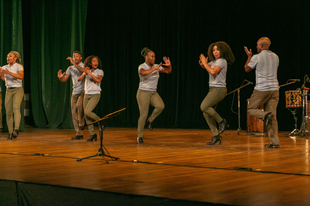 Eine Gruppe von Menschen auf einer Bühne, die jubelnd klatschen, mit Mikrofonen, Musikinstrumenten und einem Vorhang im Hintergrund.