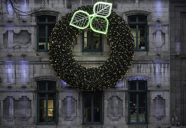 Ein festlicher Weihnachtskranz aus beleuchtetem Efeu hängt an der Seite eines Gebäudes mit Fenstern und einem Baum links daneben.