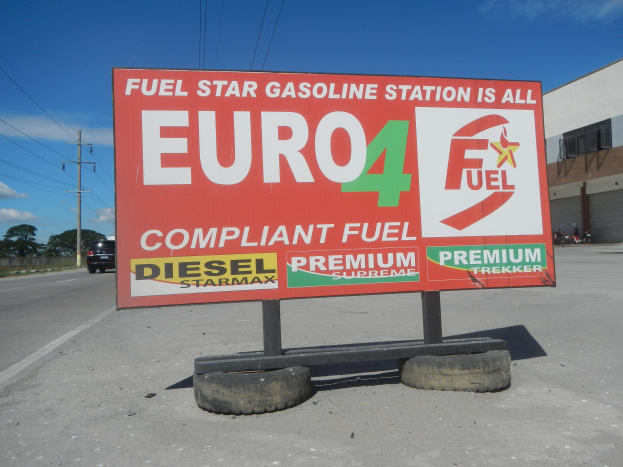 Tankstellen-Schild mit der Aufschrift "Euro4 Fuel" mit Fahrzeugen, Stromleitungen, Bäumen, einem Gebäude und Himmel im Hintergrund.