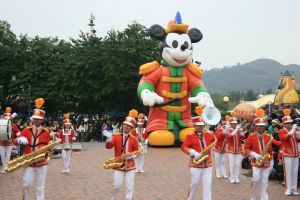 Eine Gruppe von Menschen in Kostümen spielt Musikinstrumente neben einem Mickey-Maus-Floß in einem Umzug, mit Bäumen, Gebäuden und einem klaren Himmel im Hintergrund; ein Schild mit Text ist links sichtbar.