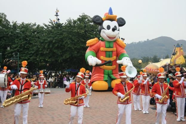 Eine Gruppe von Menschen in Kostümen spielt Musikinstrumente neben einem Mickey-Maus-Floß in einem Umzug, mit Bäumen, Gebäuden und einem klaren Himmel im Hintergrund; ein Schild mit Text ist links sichtbar.
