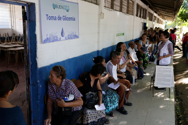 Eine Gruppe von Menschen sitzt auf Bänken vor einem Gebäude mit einer Texttafel an der Wand, einer Tür links und Bäumen im Hintergrund, die auf die Abstimmung an einer Wahllokal in Guadalajara, Mexiko, warten.