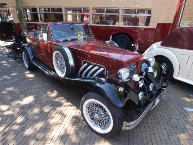 Ein Vintage-Auto neben einem Doppeldeckerbus auf einem Kopfsteinpflasterweg geparkt, mit Stühlen und einem Baum im Vordergrund und einem Gebäude mit Fenstern im Hintergrund; Menschen sind im Bus zu sehen.