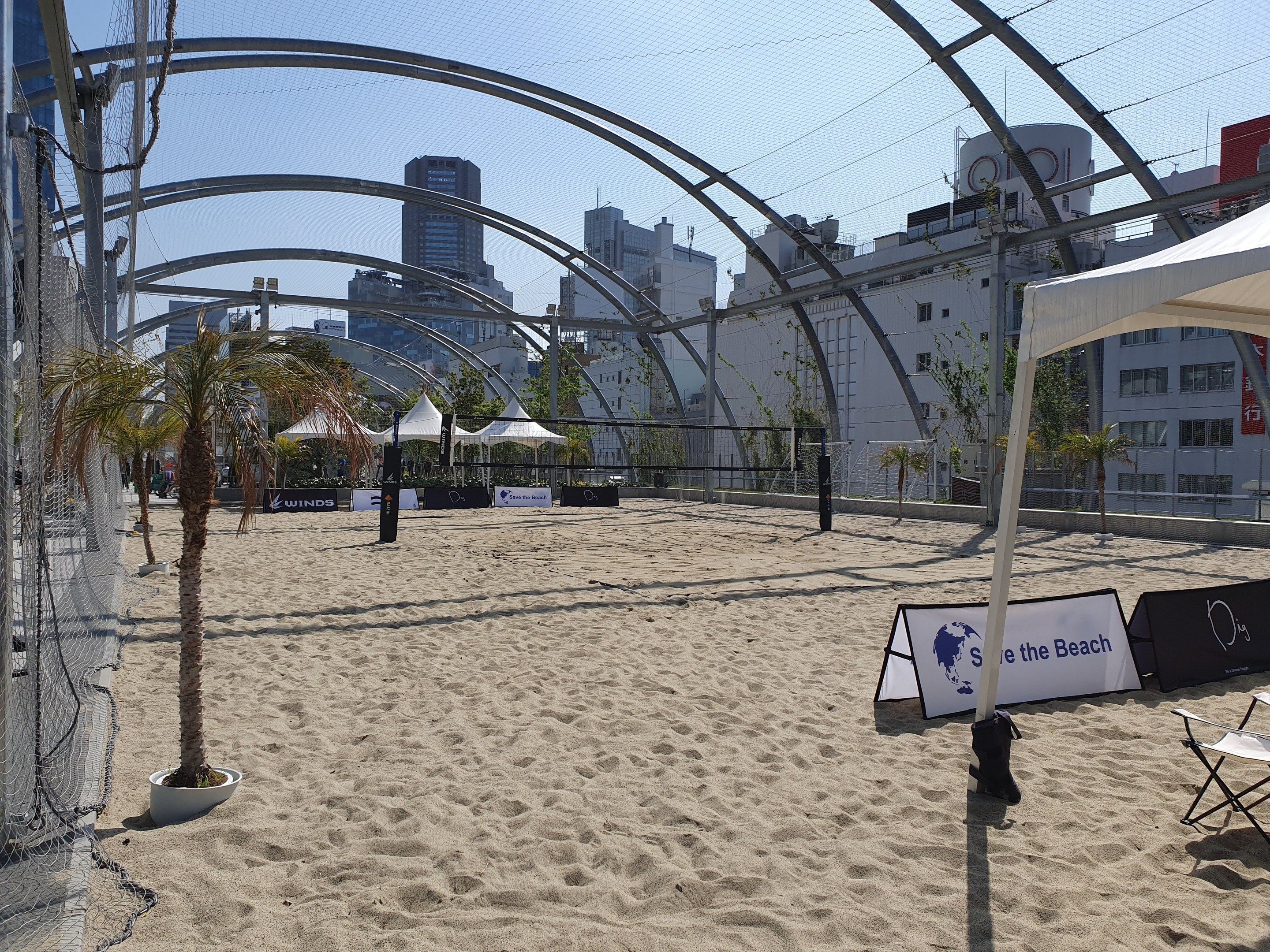 Ein Beachvolleyballfeld mit Netz und Stühlen im Sand, umgeben von Bäumen und Gebäuden, mit Bannern, Zelten und anderen verstreuten Gegenständen unter einem sichtbaren Himmel.