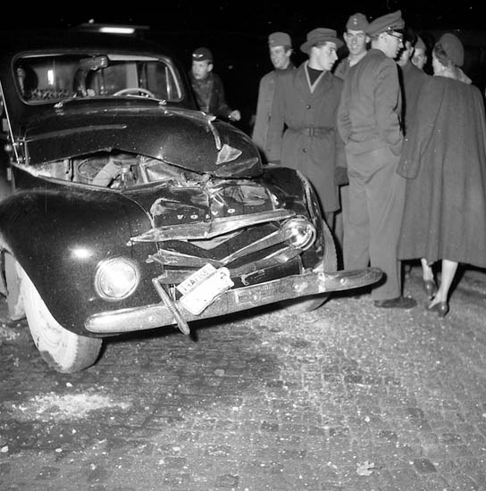 Schwarz-weißes Bild eines schwer beschädigten Autos am Straßenrand mit einer Gruppe von Menschen, die es untersuchen.