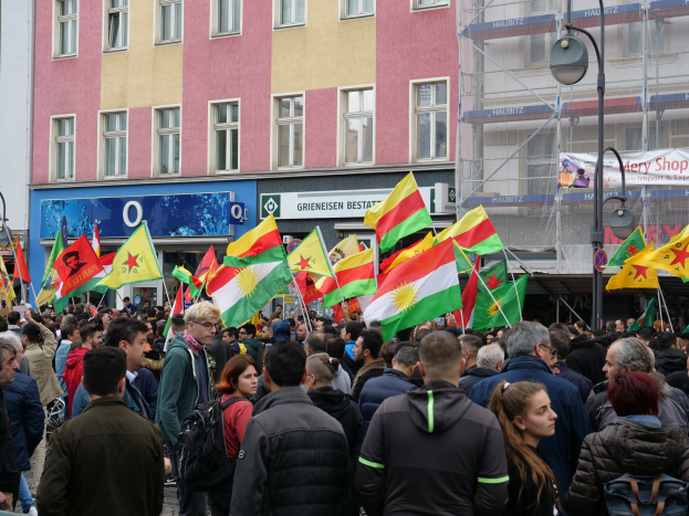 Eine große Gruppe von Menschen geht eine Straße entlang und hält kurdische Flaggen in den Händen, während Laternenmasten und Gebäude mit Fenstern die Straße säumen und Schilder mit Text im Hintergrund zu sehen sind.