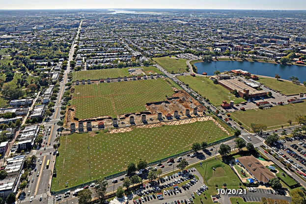 Luftaufnahme einer Stadt mit Gebäuden, Bäumen, Gras, Straßen mit Fahrzeugen, Wasser und bewölktem Himmel, die einen zentralen großen Platz zeigt, der als Park geplant ist.