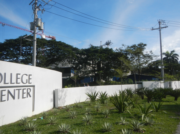 Eingang zu einem College-Center mit einem "College-Center"-Schild, umgeben von Grün, einem Weg, Strommasten, Bäumen, Gebäuden, einem Kran und einem bewölkten Himmel.