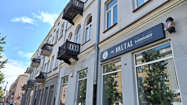 Eine Straßenansicht eines belebten Friseurladens in Berlin, Deutschland, mit Gebäuden, die Fenster, Geländer, Balkone und Schilder tragen, Bäumen, Laternenpfählen und Menschen, die auf der Straße unter einem bewölkten Himmel gehen.