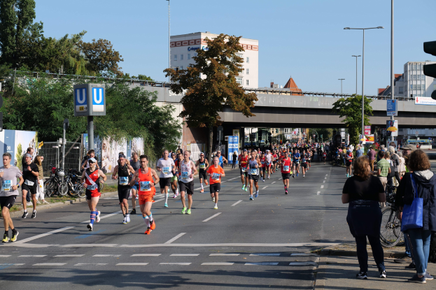 Eine Gruppe von Menschen, die auf einer von Bäumen gesäumten Straße mit Laternen, Schildern, Fahrrädern, einem Zaun, Gras, einer Brücke, Gebäuden und einem klaren blauen Himmel laufen.
