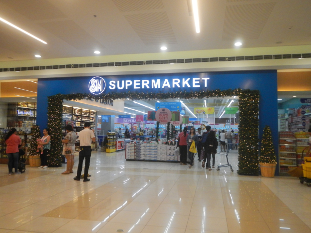 Menschen vor einem Supermarkt mit Einkaufstaschen, einige mit einem Einkaufswagen, Topfpflanze, Regalen, Schildern und Deckenbeleuchtung.