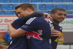 Zwei Fussballspieler umarmen sich auf einem Feld, mit einer Hand sichtbar links und im Hintergrund Menschen, Schilder mit Text und blaue Stühle.