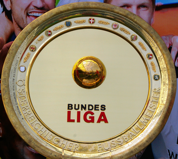 Ein Mann hält einen goldenen Schild mit dem Wort "Bundesliga" darauf, mit einem Banner im Hintergrund mit drei Personen.